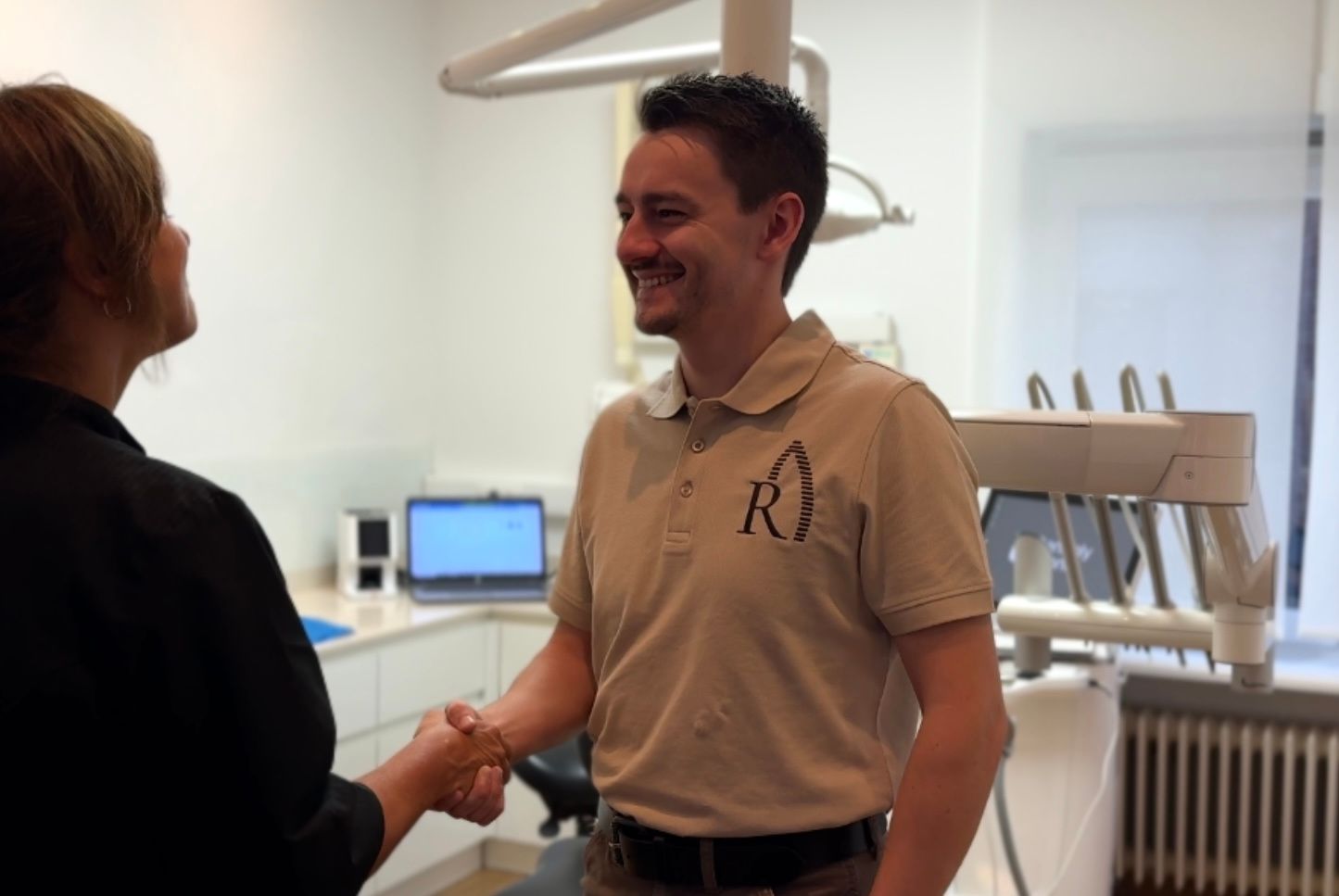 Een tandtechnieker in een Retzius-polo geeft een tandarts een hand in de behandelkamer, naast de stoel.
